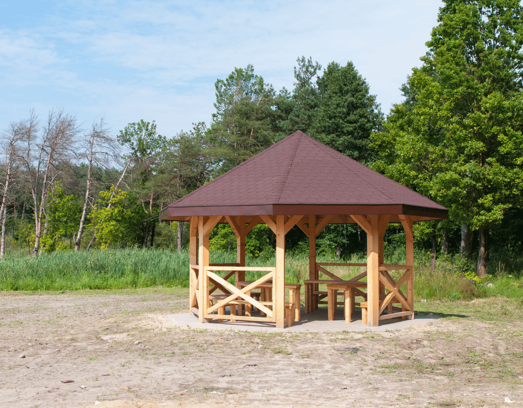 Wood-framed gazebo with a reddish-brown shingle roof near a wooded area
