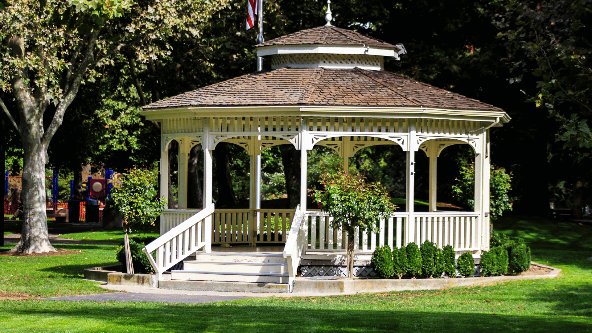 White Victorian-style gazebo with a shingled roof and an American flag in a park