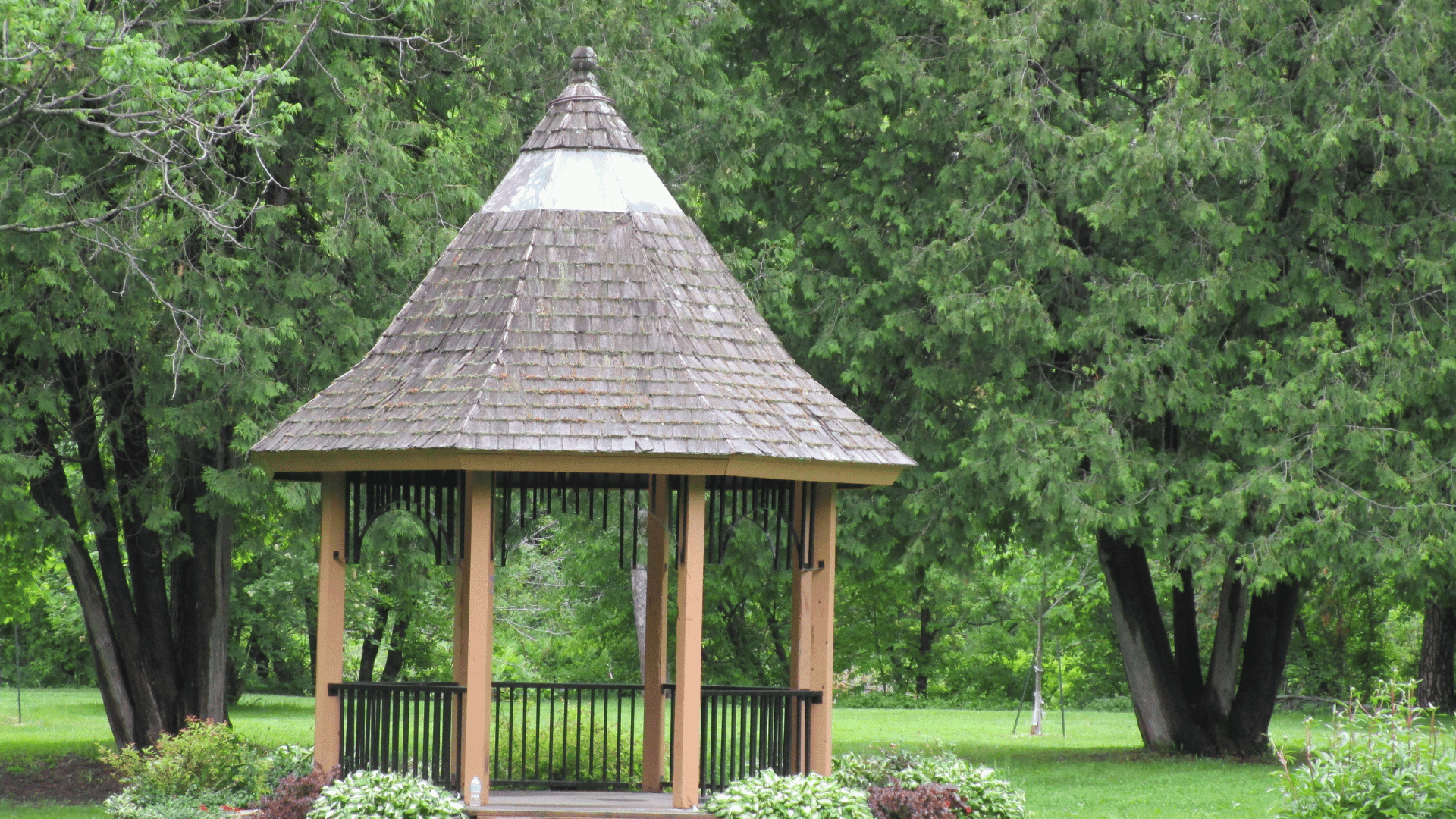 Wood-framed pavilion with a shingled roof surrounded by mature trees